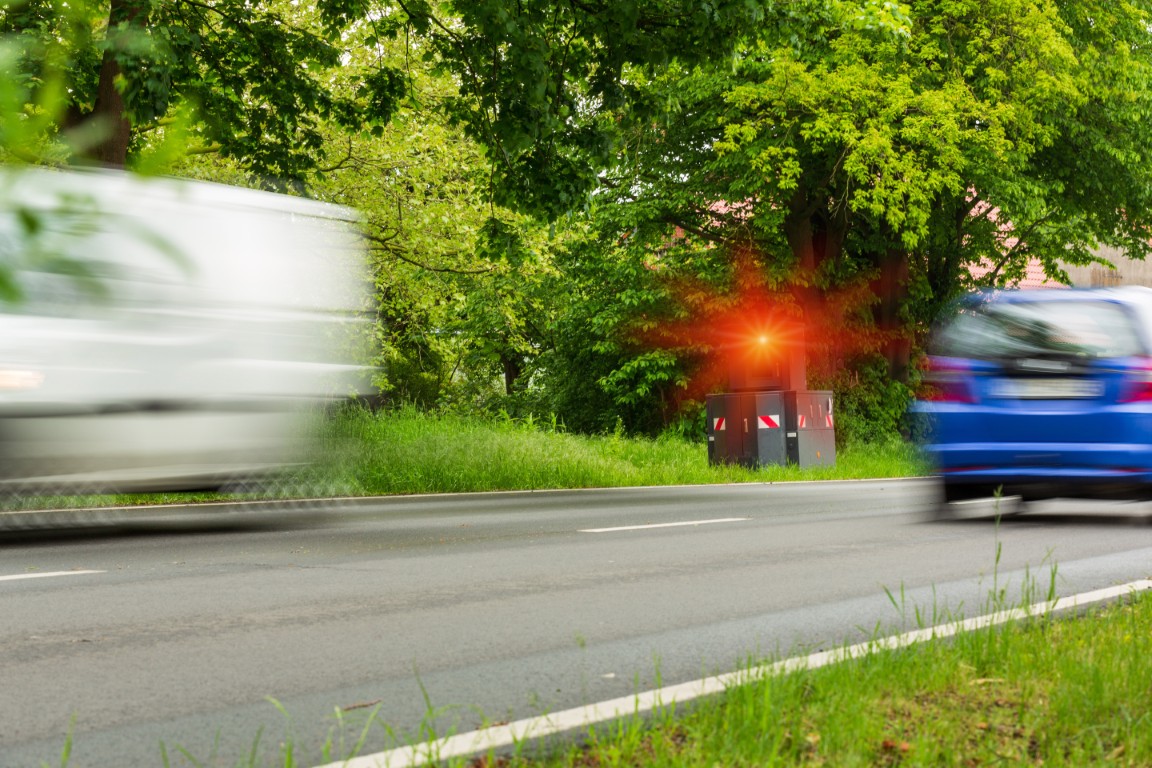 Ein weißer Lieferwagen und ein blaues Auto rasen eine begrünte Straße in der Gemeinde Moorenweis hinunter und passieren einen Blitzer, der rot leuchtet.