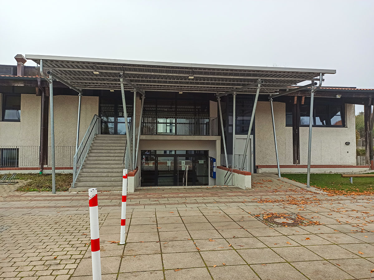 Ein moderner Gebäudeeingang in der Gemeinde Moorenweis mit einem großen Metallvordach, breiten Stufen auf der linken Seite, Glastüren und weißen Wänden. Die gepflasterte Fläche davor ist mit rot-weißen Pfählen und verstreutem Herbstlaub versehen.