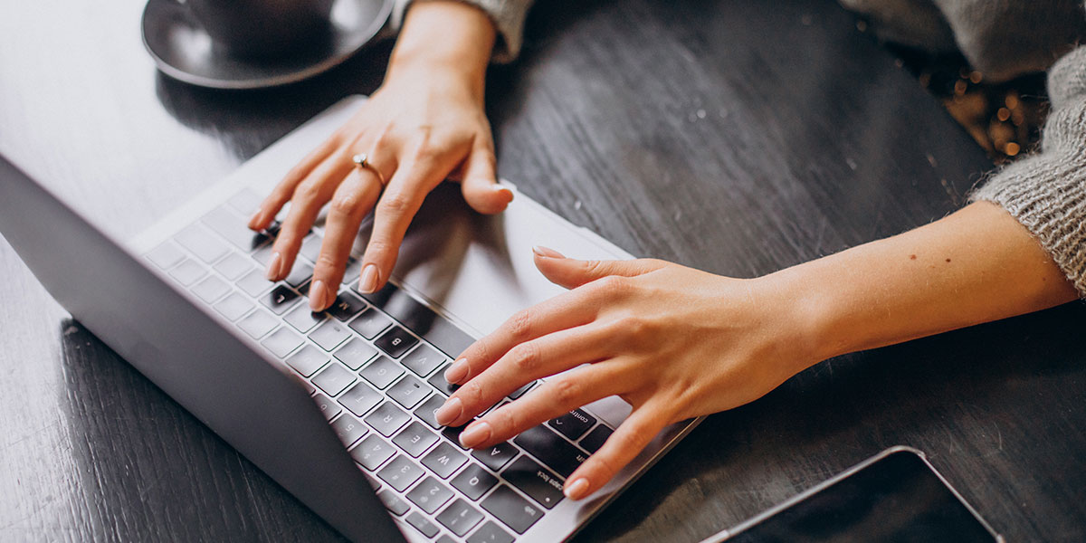 Nahaufnahme der Hände einer Person, die auf einer Laptoptastatur an einem dunklen Holztisch in der Gemeinde Moorenweis tippt. Im Hintergrund sind eine Tasse mit Untertasse und ein Smartphone zu sehen. Die Person trägt einen grauen Pullover.