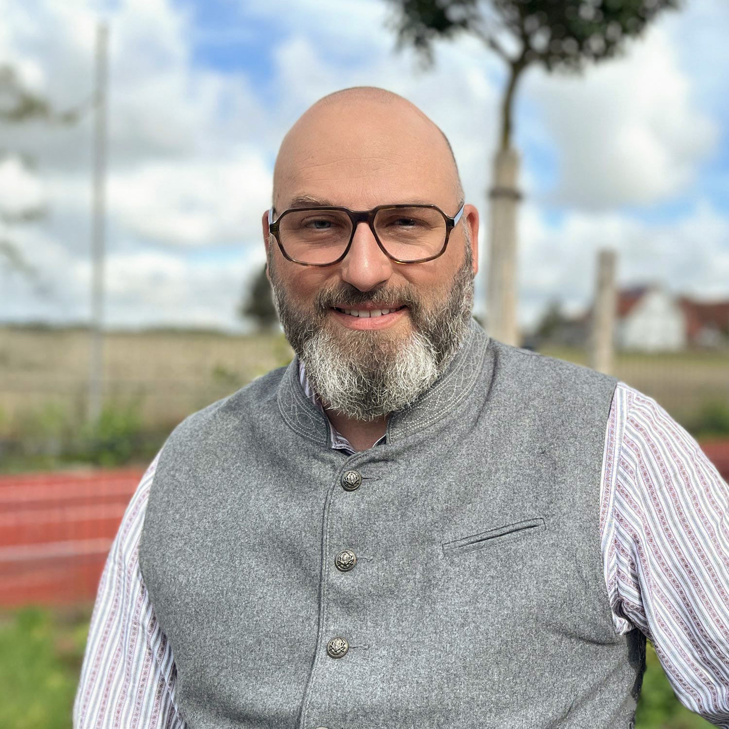 Ein lächelnder Mann mit Glatze, Brille und grauem Bart trägt eine graue geknöpfte Weste über einem gestreiften Hemd. Er steht im Freien in der Gemeinde Moorenweis mit einem unscharfen Hintergrund aus einem Zaun, Bäumen und einem bewölkten Himmel.