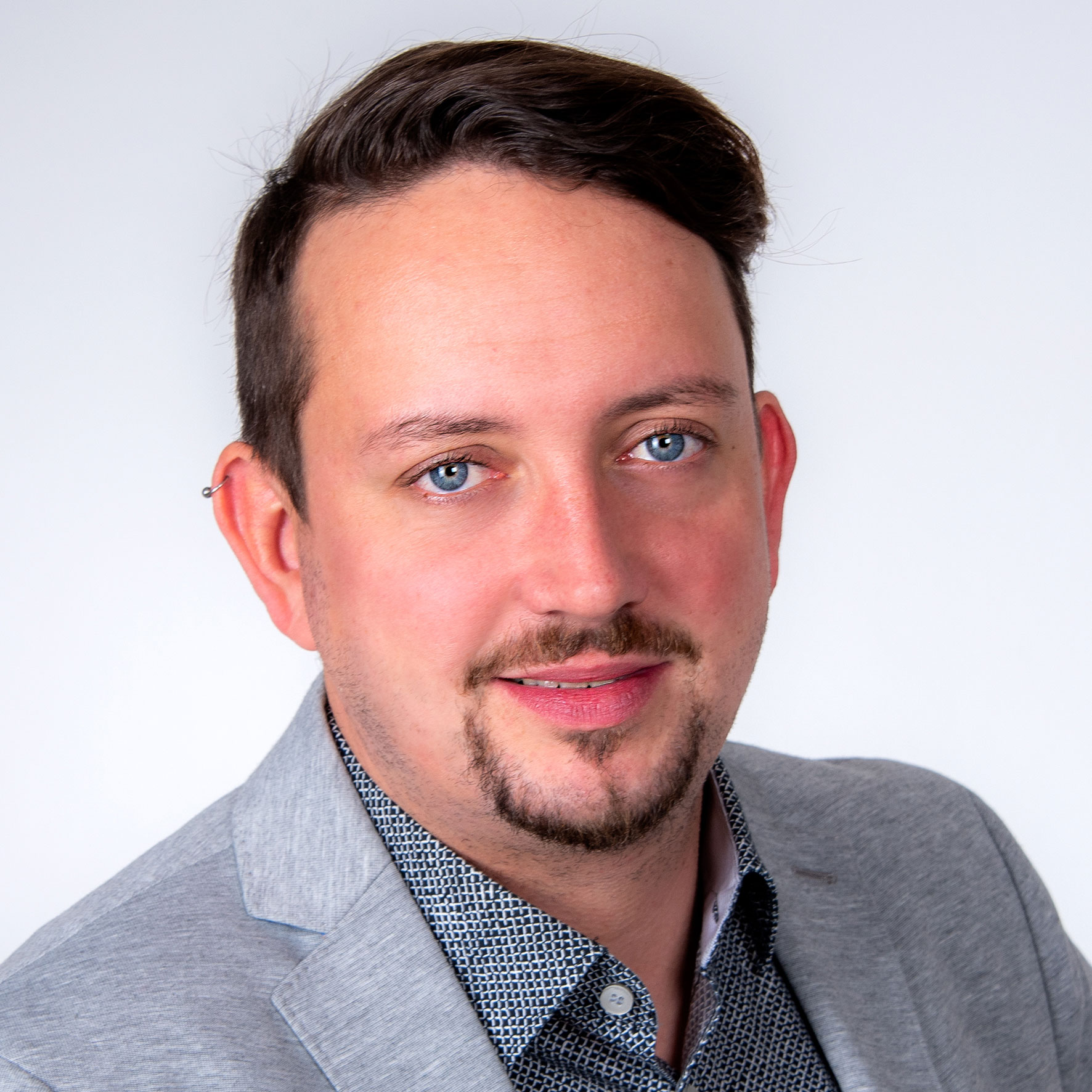 Michael Leib Ein Mann mit kurzen braunen Haaren, einem gestutzten Bart und blauen Augen trägt einen hellgrauen Blazer über einem gemusterten Hemd. Er posiert vor einem schlichten, hellen Hintergrund und lächelt leicht.