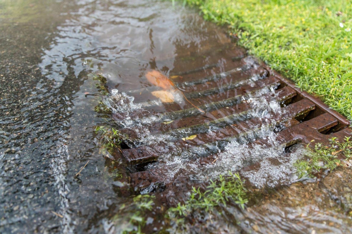 Regenwasser fließt über ein Metallgitter am Rande einer Grasfläche und eines Gehwegs, wobei das Wasser Laub und Schmutz mit sich führt, während es in den Abfluss gelangt.
