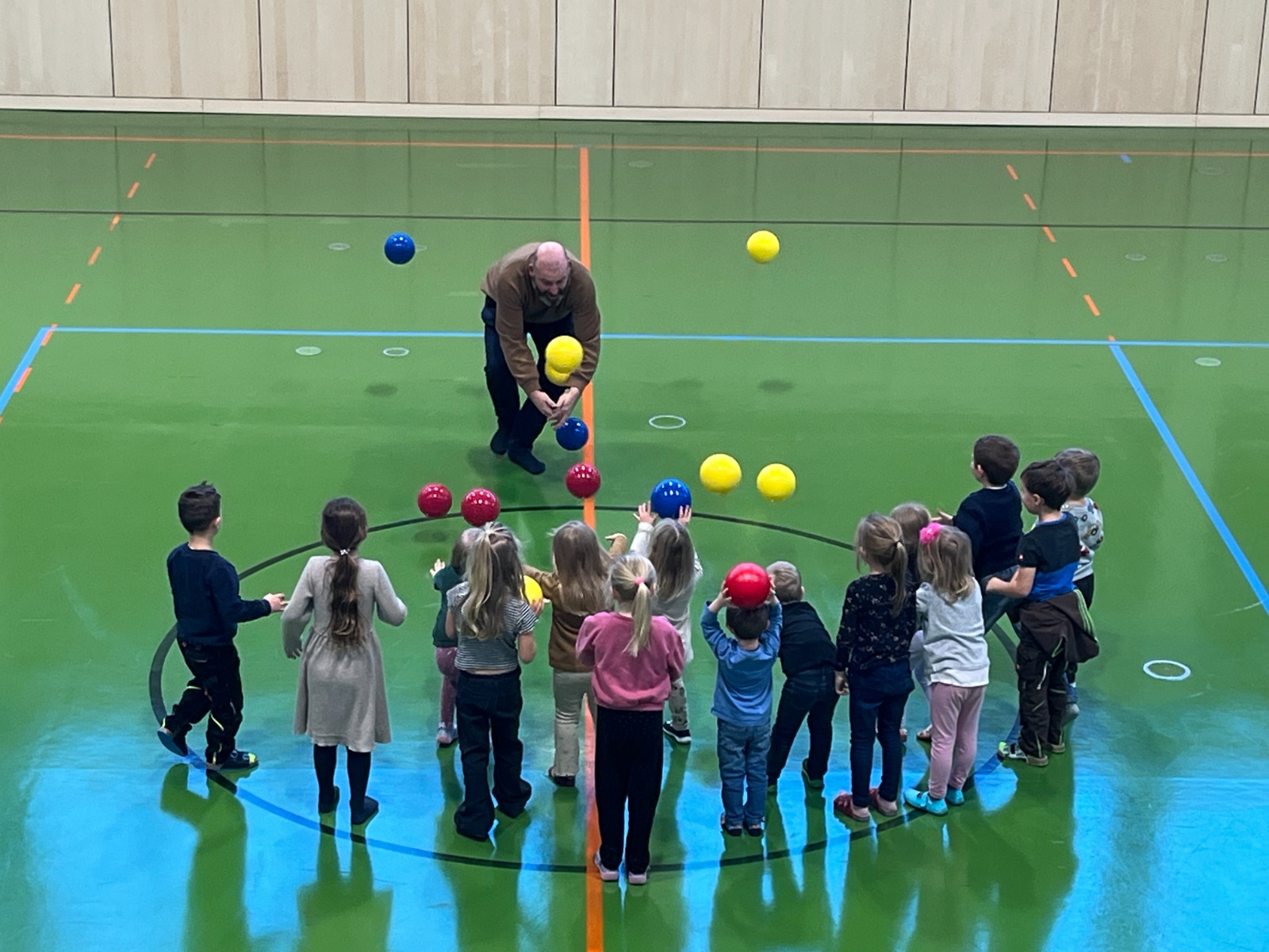 Eine Gruppe kleiner Kinder steht in einer Turnhalle im Kreis um einen Erwachsenen. Der Erwachsene jongliert mit mehreren bunten Bällen, während die Kinder zusehen und mitmachen, einige halten Bälle. Der Boden ist grün mit blauen und orangefarbenen Sportplatzlinien.