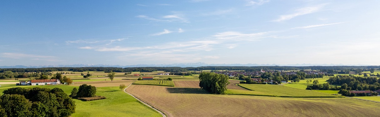 Eine weite Luftaufnahme einer ländlichen Landschaft mit grünen Feldern, verstreuten Bäumen, Bauernhäusern und Dörfern unter einem blauen Himmel mit leichten Wolken; am Horizont sind ferne Berge zu sehen.