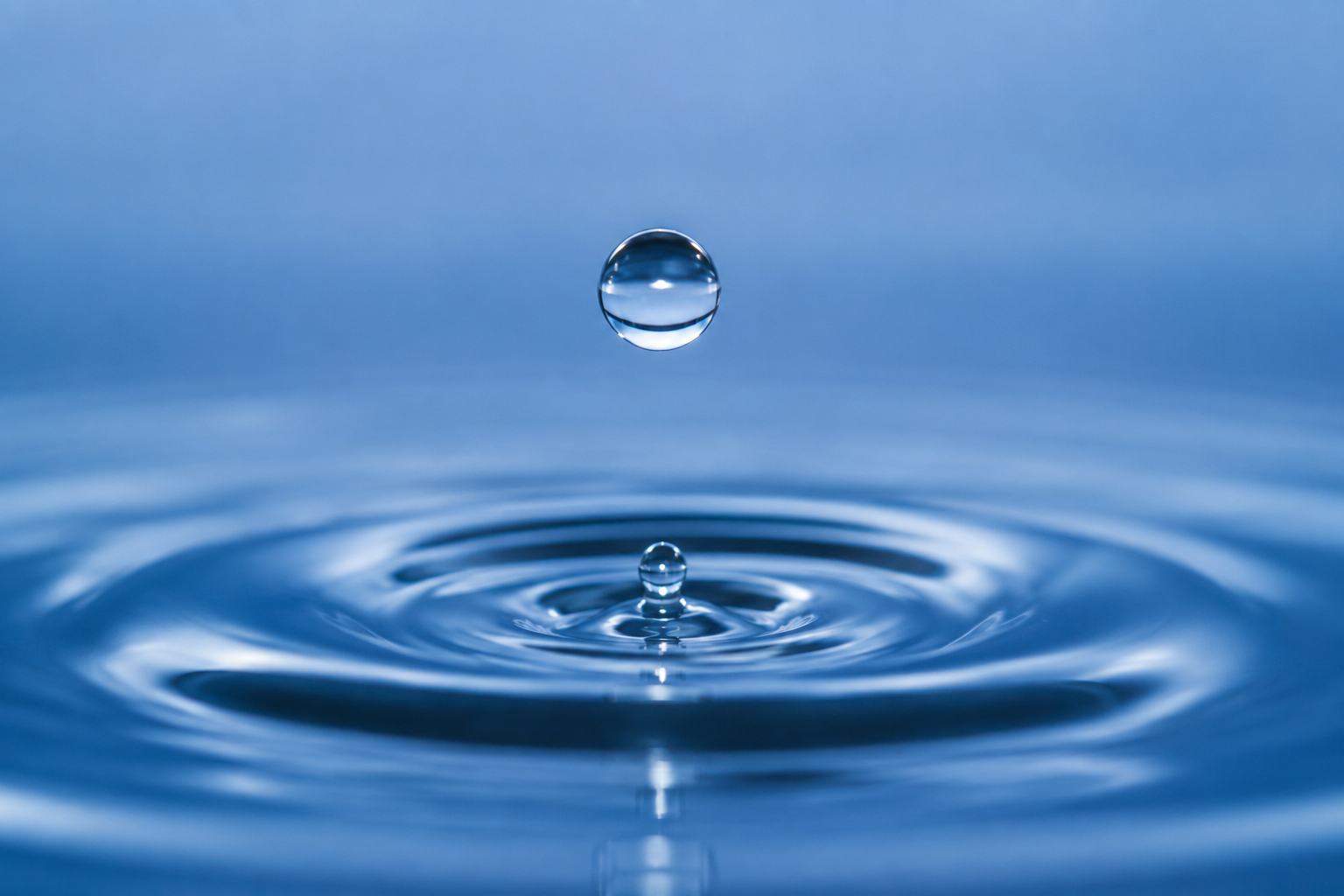 Eine Nahaufnahme eines einzelnen Wassertropfens, der über eine ruhige Wasseroberfläche fällt und dabei kreisförmige Wellen erzeugt. Das Bild zeigt kühle Blautöne und eine glatte, reflektierende Textur.