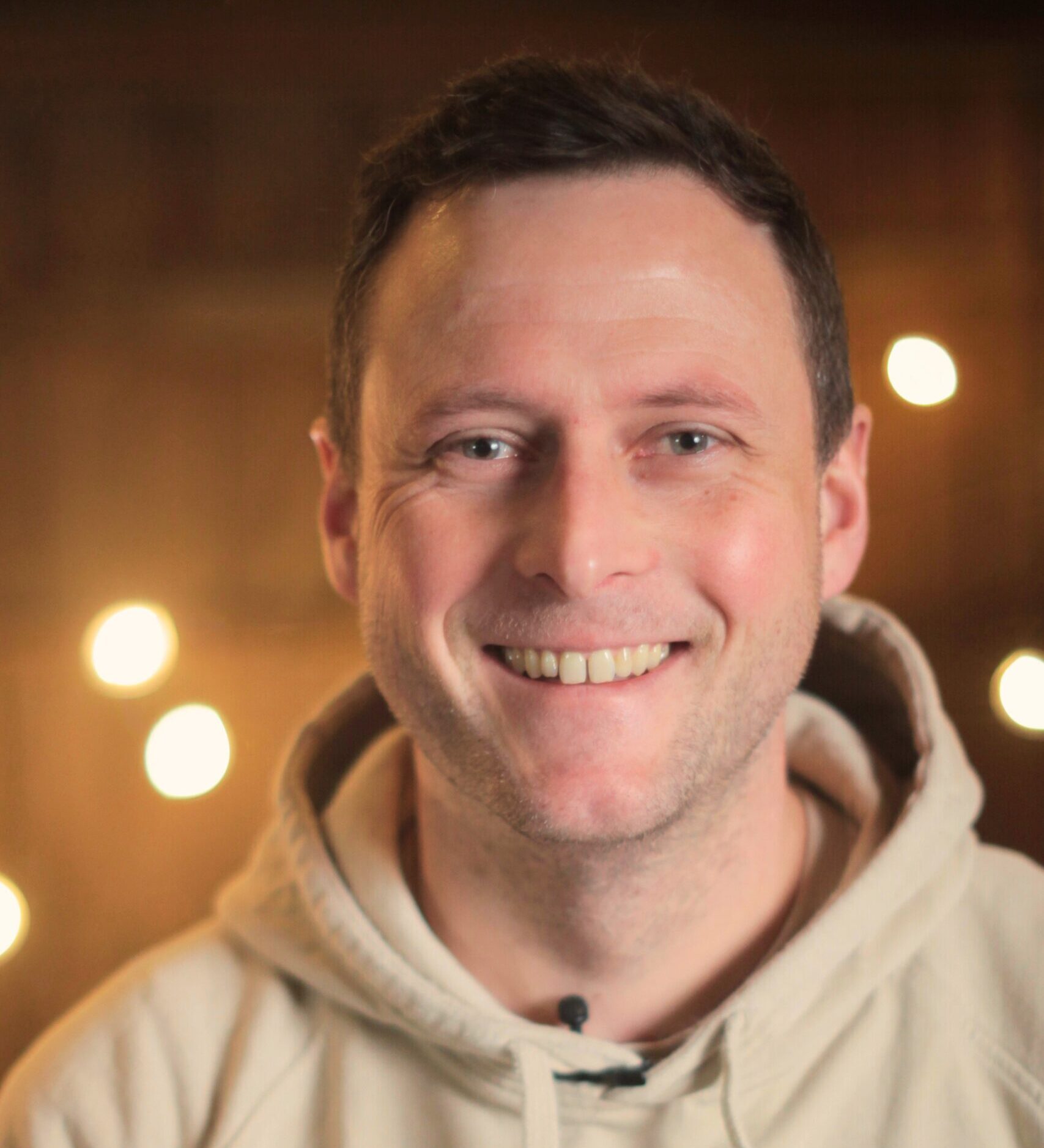 Ein Mann mit kurzen braunen Haaren lächelt in die Kamera; er trägt einen beigen Kapuzenpulli. Im Hintergrund leuchten warme, unscharfe Lichterketten und sorgen für eine gemütliche Atmosphäre.