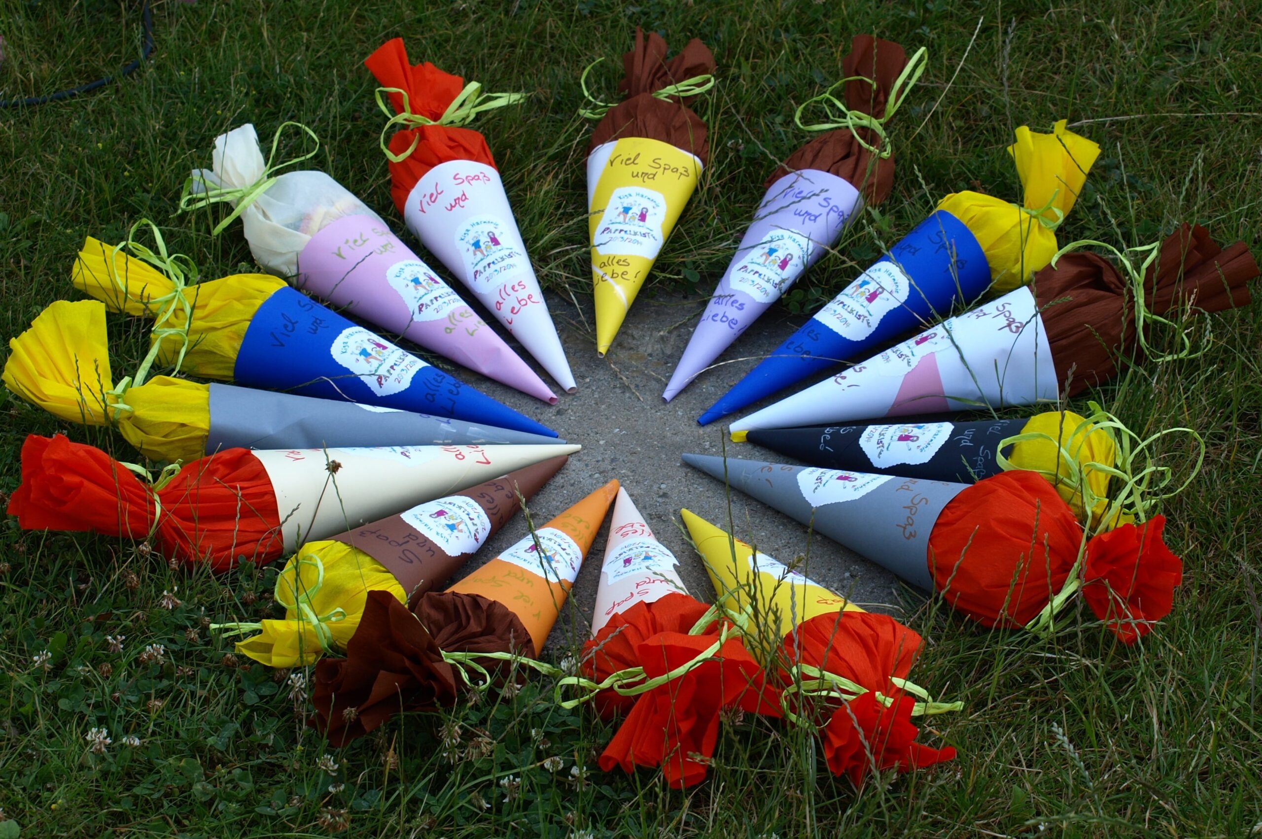 Bunte Papierkegel mit handgeschriebenen Notizen und Seidenpapierhauben, die im Kreis auf dem Rasen angeordnet und jeweils mit einem Band zusammengebunden sind; diese werden in Deutschland üblicherweise zum ersten Schultag der Kinder verwendet.