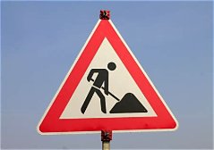 Dreieckiges Verkehrszeichen mit rotem Rand und der schwarzen Silhouette einer Person, die gräbt, das auf bevorstehende Straßenbauarbeiten hinweist, vor dem Hintergrund eines strahlend blauen Himmels.